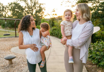 Two young mothers talk happily as they hold their toddler daughters on their hips and walk through the park at sunset. One of the women is eight months pregnant.